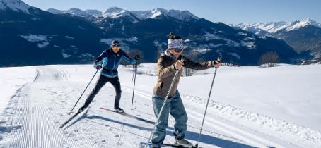 Couple de fondeurs sur une piste enneigée avec vue dégagée sur une chaine de montagne.